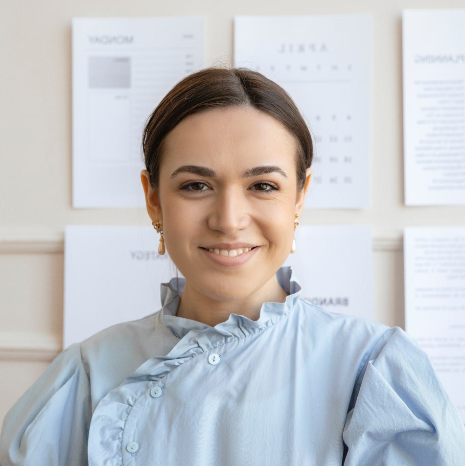 Business consultant smiling in a professional setting, wearing a light blue blouse, with planning documents and schedules visible in the background, emphasizing a focus on organization and efficiency in project management.