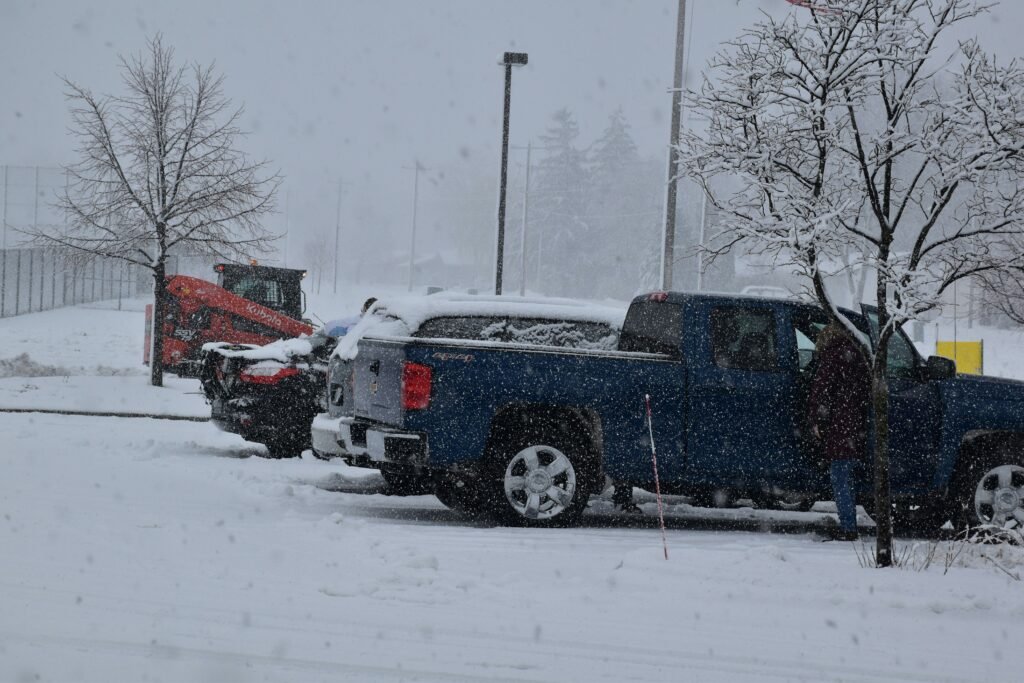 Blue pickup truck in snowy parking lot, snow removal equipment in background, winter storm conditions affecting visibility and access, illustrating challenges of commercial snow management.